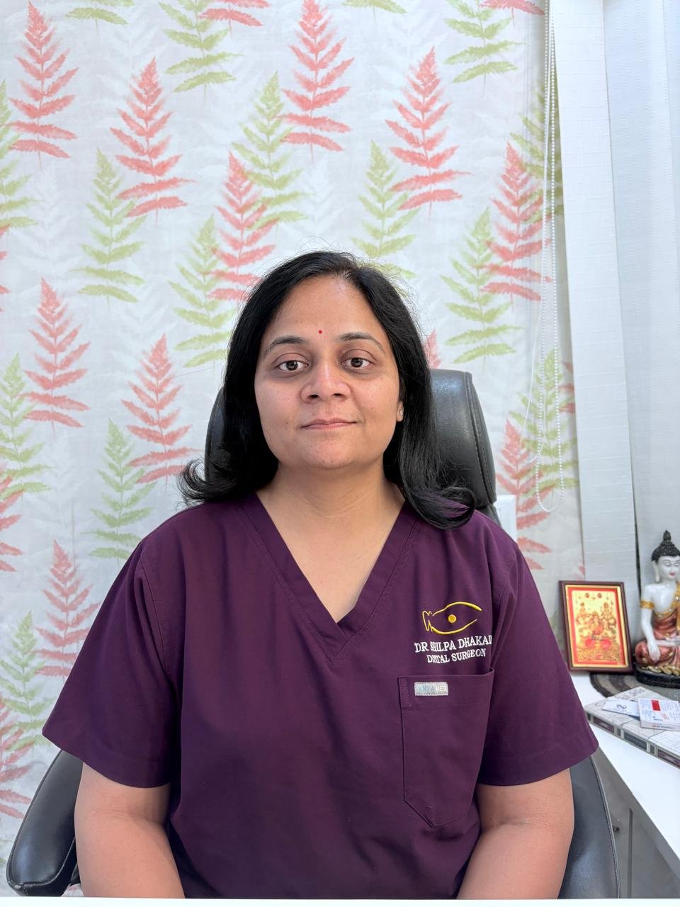 Dentist in clinic wearing professional scrubs, seated in a clean consultation room.
