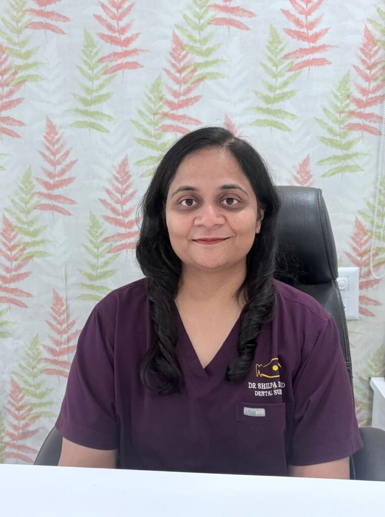 Female dentist seated in clinic wearing maroon dental scrubs.